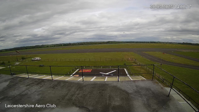 A view from a webcam overlooking the Leicester Aero Club, showing a grassy area with a helicopter landing pad painted with a white helicopter silhouette. In the background, there is an empty taxiway and runway with some distant green fields and a cloudy sky overhead. There is a small white vehicle parked near a flagpole to the left, and a few small structures and signs are also visible. The timestamp in the top right corner reads "30-05-2025 14:00:17".