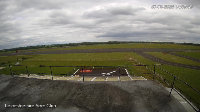 A view from a webcam at Leicesterhire Aero Club showing an overcast sky with scattered clouds, a grassy airfield with runways in the distance, a windsock on the left side, a helipad with a white "H" in the foreground, and a railing along the edge of a balcony or viewing platform. The timestamp indicates the date as May 30, 2025, at 14:59:59.
