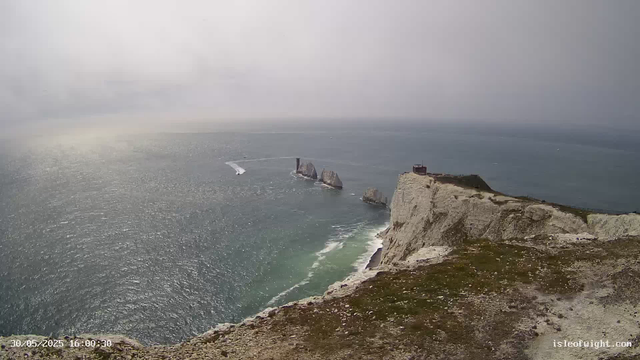A view from a cliff overlooking the ocean with tall sea stacks in the distance, some with structures on top, under a cloudy sky. The foreground shows rocky and grassy terrain at the edge of the cliff.