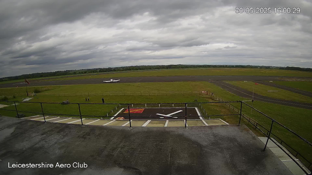 An overcast sky looms over a small airport runway, with a single small airplane parked in the distance. In the foreground, there is a helipad marked with a large white "H" on a black background, bordered by a fence. A few people are standing on the grass near the runway, and a windsock is visible to the left. The image is labeled "Leicestershire Aero Club" in the bottom left corner, and a timestamp in the top right corner reads "30-05-2025 16:00:29."