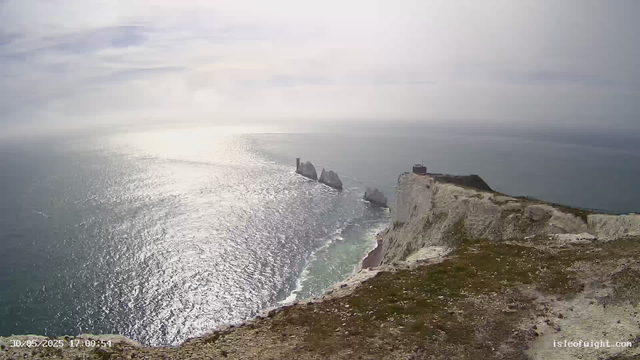 A view of steep white chalk cliffs overlooking the ocean. The sea is shimmering with sunlight, creating a bright reflection on the water. A few large rock formations are visible offshore, with one having a small lighthouse or tower and another with a building on top. The sky is partly cloudy, with soft, diffuse light illuminating the scene.