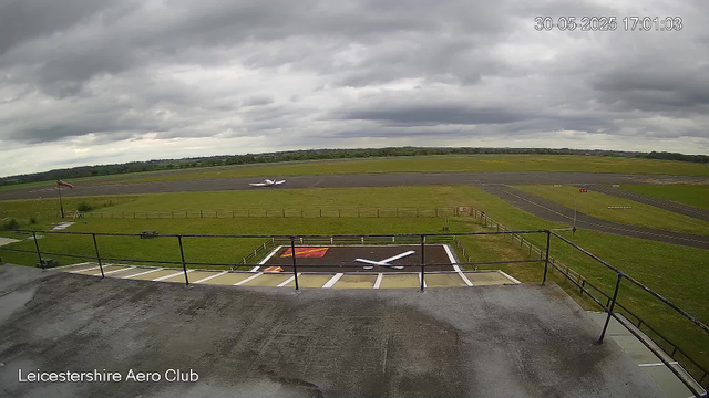 A view from a webcam at Leicesterhshire Aero Club showing an overcast sky with dark clouds. In the foreground, there is a small helipad with a white helicopter parked on it. A windsock is visible to the left, nearby a flagpole with a flag. The airfield's runway extends into the distance, surrounded by green grass and fields, with a distant horizon and a line of trees under the cloudy sky. The timestamp in the top right corner reads "30-05-2025 17:01:03."