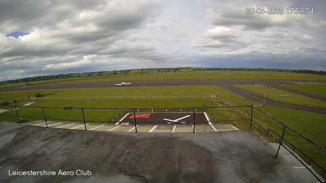 A panoramic view of an airfield taken from a webcam. In the foreground, there is a rooftop with a railing, and a helicopter landing pad with a large white "H" on it. Beyond the rooftop, there's a small plane taxiing on the runway, with green fields and tarmac stretches in the background. The sky is mostly cloudy with patches of blue, and the timestamp in the top right corner reads "30-05-2025 17:59:54." The bottom left corner has the text "Leicestershire Aero Club."