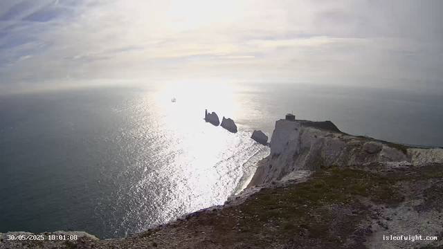 A panoramic view of white chalk cliffs meeting the ocean under a partly cloudy sky. The sunlight reflects off the water, creating a shimmering effect. In the distance, a sailboat is visible on the horizon. The cliff edges include a small building and a lighthouse-like structure. The foreground shows a rocky and grassy terrain. The timestamp indicates the image was captured on May 30, 2025, at 18:01.