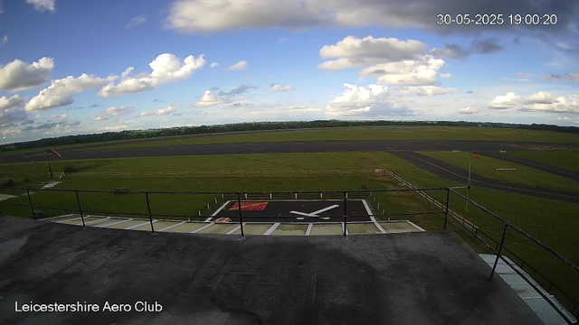A view from a webcam at Leicester Airport shows a clear day with a partly cloudy sky. In the foreground, there is a helicopter landing pad with a large white "X" marked in the center. Beyond the helicopter pad, there is a grassy area and a runway extending into the distance. The landscape is flat with some trees visible on the horizon. The top right corner displays the date and time as 30-05-2025 19:00:20. The bottom left corner contains the text "Leicestershire Aero Club."