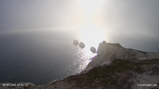 A view of white chalk cliffs along a coastline with a small building or lighthouse on top, overlooking the sea. The sun is low on the horizon, creating bright reflections on the water and casting a glow over the scene. Rocks and the rugged cliff edge are visible in the foreground. The image is timestamped "30/05/2025 19:01:11" and includes the website "isleofwight.com" in the bottom right corner.