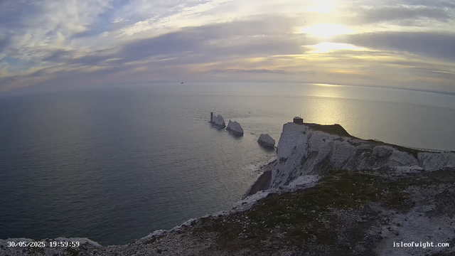 A view of white chalk cliffs overlooking the sea at sunset, with a small building and a fence on the edge of the cliff. The sky is partly cloudy with the sun low on the horizon, casting a golden reflection on the water. In the distance, two large sea stacks with a small lighthouse between them are visible in the calm ocean. The foreground shows the rocky edge of the cliff.