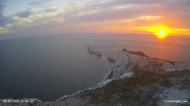 A sunset over white chalk cliffs on the coast, with the sea stretching out to the horizon. The sky is partly cloudy with shades of pink, purple, and orange. There are three of the iconic chalk stacks in the water, with one featuring a lighthouse or tower on top, and a small building or lookout on the highest point of the cliffs. The image is timestamped "30/05/2025 21:00:16" and has the website "isleofwight.com" in the bottom corner.