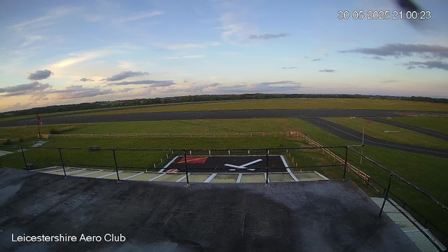 A view from a webcam at Leicesterhire Aero Club shows a runway and surrounding grassy areas under a partly cloudy sky during the early evening. The image includes a large clock logo on the ground, resembling an airplane propeller, and a small red and white sign near the runway. The timestamp in the top right corner reads "30-05-2025 21:00:23."