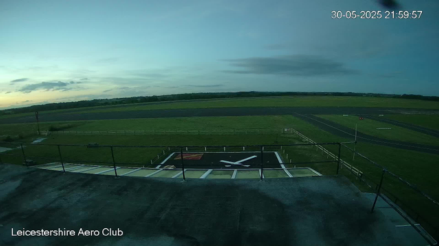 An empty helicopter landing pad with a large "X" symbol is visible in the foreground, surrounded by a fenced area. Beyond the pad, there are green fields and a runway extending into the distance. The sky is partly cloudy with a fading sunset, creating a calm evening atmosphere. The image has a timestamp in the upper right corner showing the date and time as 30-05-2025 at 21:59:57, and a label in the bottom left corner that reads "Leicestershire Aero Club".
