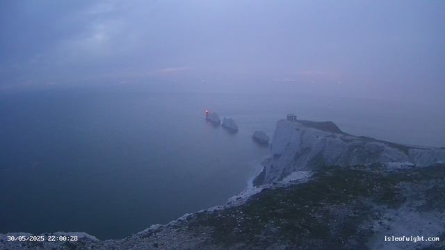 A view of steep white chalk cliffs extending into the calm sea at dusk or dawn, with a lighthouse on the edge of the cliffs and several large rocks emerging from the water nearby. The sky is overcast with a bluish hue, and there are faint lights visible in the distant background across the water. The timestamp in the bottom left corner indicates the date and time as May 30, 2025, at 22:00:28. The website "isleofwight.com" is displayed in the bottom right corner.