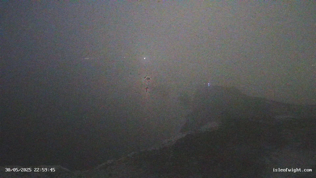 A dark, nighttime view of a coastal area with the ocean in the foreground. The sky above is cloudy with visible stars and a few aircraft or satellite lights. The shoreline is faintly visible in the lower part of the image, and a mountain or hill silhouette is seen in the background. The timestamp shows "30/05/2025 22:59:45" and the URL in the corner indicates a webcam from isleofwight.com.