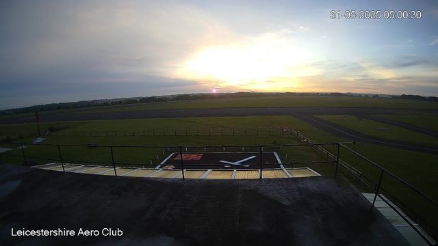 A sunrise over an airfield with a large clock on the ground and a railing in the foreground. The sky is partly cloudy, and the horizon shows grassy fields and distant trees. The timestamp in the top right corner reads "31-05-2025 05:00:30," and the bottom left corner has the text "Leicestershire Aero Club."