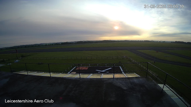 A view from a webcam at Leicestershire Aero Club showing an open area with a grassy field, a runway in the distance, and a large black clock on a platform in the foreground. The sky is partly cloudy with the sun low on the horizon, suggesting early morning or late evening. The timestamp in the top right corner reads "31-05-2025 06:00:10."