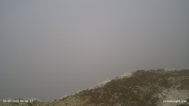 A foggy landscape showing the top of a rocky hill or mountain. The sky is overcast and visibility is low due to thick fog. The terrain appears rugged with patches of dirt and rocks. The image has a timestamp at the bottom left corner reading "31/05/2025 06:00:47" and a website URL "isleofwight.com" at the bottom right.