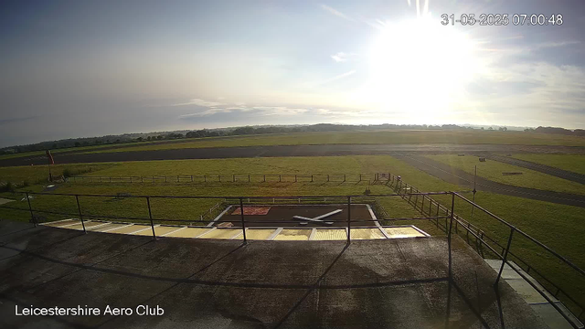 A wide view of an outdoor area at Leicesterhire Aero Club during sunrise or sunset, with a vast grassy field and open skies. The foreground shows part of a rooftop or balcony with metal railings and a small fenced-off section that appears to be a tennis or sports court. The sky is partly cloudy with the sun shining brightly, casting strong light over the scene. The timestamp indicates the image was taken on May 31, 2025, at 7:00 AM.