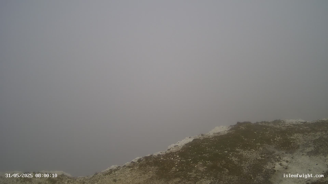 A foggy, overcast sky obscures the view of the landscape. In the foreground, there is a rocky, grassy hillside with patches of white, possibly snow or frost. No other objects or features are visible through the dense fog. A timestamp at the bottom left shows the date and time as "31/05/2025 08:00:10," and the bottom right has the website "isleofwight.com."