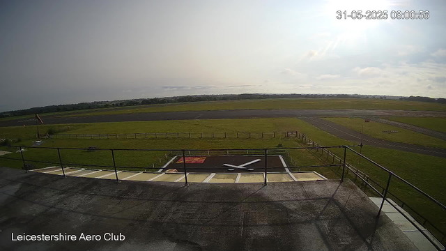 An empty outdoor airfield with a helipad in the foreground, situated within the Leicestershire Aero Club. The helipad has a large "H" marked on it, and there is a red doormat nearby. Surrounding the airfield are grassy fields, a runway, and a clear sky with the sun shining brightly. The timestamp in the upper right corner indicates the date and time as 31-05-2025, 08:00:56.