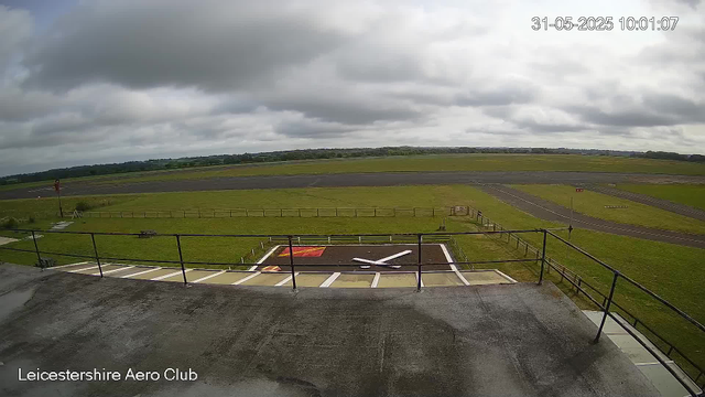 An empty aircraft runway under a cloudy sky, with a helicopter landing pad marked in red and yellow in the foreground. The scene is overcast, and the surrounding grassy area is green. The timestamp in the top right corner shows "31-05-2025 10:01:07," and there is a label at the bottom left that reads "Leicestershire Aero Club."