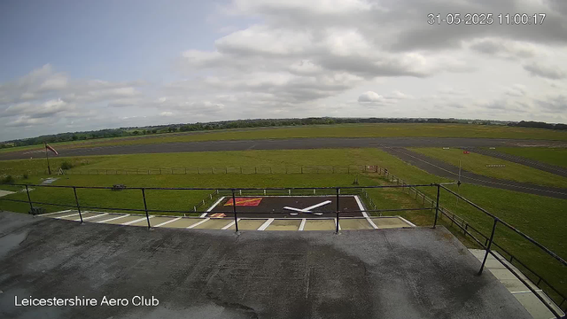 A view from a webcam at Leicestershire Aero Club showing an open grassy area with a small helipad marked with a white helicopter symbol in the foreground. Beyond the helipad, there is a green field with a flagpole on the left side. The background features a runway and taxiways, with a cloudy sky overhead. The scene appears calm with no visible aircraft or people.