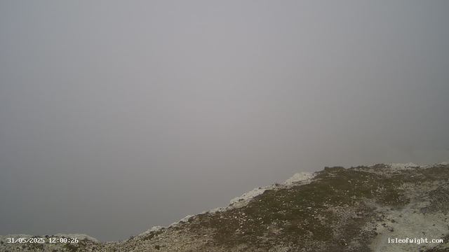 A foggy scene on a mountain ridge with a rocky, uneven surface near the bottom right corner. The sky is completely obscured by dense fog, creating a gray, uniform backdrop. The image has a timestamp in the bottom left corner, indicating it was taken on May 31, 2025, at 12:00:26.