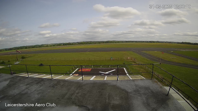 A wide view of Leicester Aero Club shows the airfield with runways, grassy areas, and some small structures. The sky is partly cloudy with scattered clouds and sunlight. In the foreground, there is a large black and white airplane sign painted on a section of the tarmac, surrounded by a railing and parking spaces. A windsock is visible on the left side, and a few people are walking on the grass. The timestamp in the upper right corner indicates the date and time as 31st May 2025 at 12:00:10.