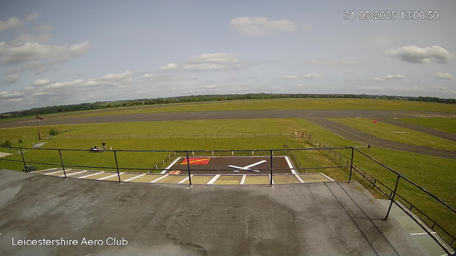 A view from a webcam at Leicestershire Aero Club showing an outdoor helicopter landing pad with a white helicopter silhouette painted on the ground. There is a grassy area with a few small structures, a person standing near a small vehicle, and a windsock on a pole. In the distance, there is a runway with runway markings, and the landscape extends to the horizon with a partly cloudy sky overhead. The timestamp reads 31-05-2025 13:00:50.
