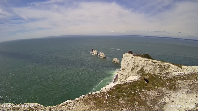 A high-angle view of a coastal scene with white chalk cliffs extending into the sea. The ocean is calm and has a gradient of green and blue hues. In the distance, there is a lighthouse standing on a small rocky outcrop, with a boat nearby creating a wake. The sky is partly cloudy with patches of blue. The foreground shows the edge of the cliff with some grass and rocks.
