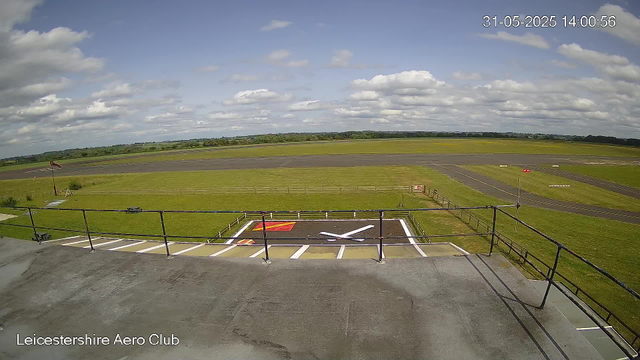 An empty tarmac runway with a grassy area and a windsock on a pole to the left. The sky is partly cloudy with white clouds scattered across a blue sky. There are painted markings and barriers on the ground in front, with the text "Leicestershire Aero Club" in the bottom left corner. The timestamp in the top right corner shows 31-05-2025 14:00:56.