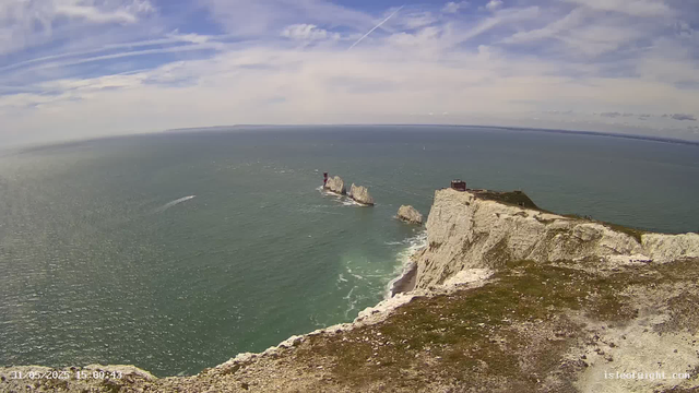A panoramic view of a coastal cliff with white chalk formations extending into the sea. There is a small lighthouse on one of the rocks in the water and a building on top of the cliff. The sky is partly cloudy with streaks of clouds and contrails from airplanes. The water below is calm with gentle waves, and a few boats are visible in the distance. The image was captured during the daytime.