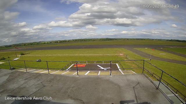 A view from a webcam at Leicestershire Aero Club shows a clear sky with scattered clouds. In the foreground, there is a small fenced area with painted markings on the ground, including a white circle with a line and some red and white markings. There is a flagpole with a windsock on the left side. An empty cart is parked nearby, and the surrounding area consists of grassy fields and a paved runway extending into the distance. The scene appears to be taken during the daytime on May 31, 2025, at 4:00 PM.