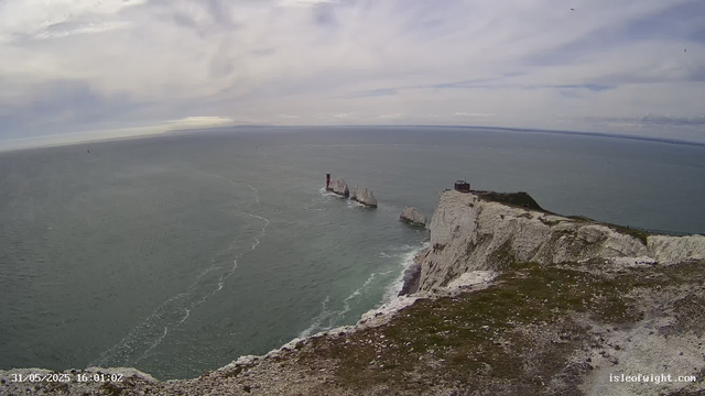 A panoramic view of a coastal landscape with tall white chalk cliffs extending into the sea. The cliffs have some dark structures on top, possibly small buildings or observation points. In the water near the cliffs, there are several large rocks or sea stacks, with one notable red lighthouse on a rock formation. The sea is calm with gentle waves, and the sky above is partly cloudy with patches of blue. The image appears to be captured from a webcam, with a timestamp in the bottom left corner reading "31/05/2025 16:01:02."