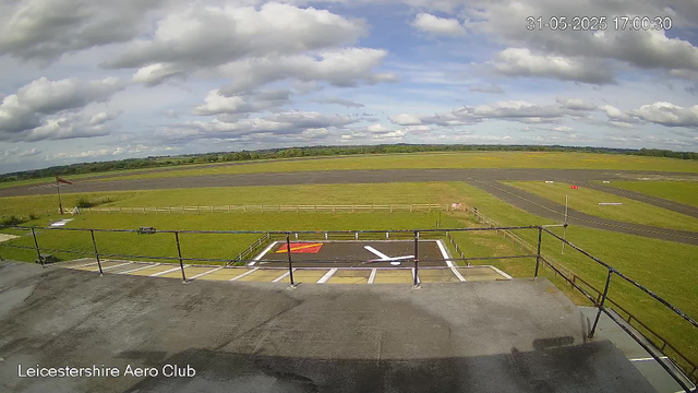 An image of an airport runway taken by a webcam shows a clear, partly cloudy sky with scattered clouds and a vast green field stretching into the distance. In the foreground, there is a small parking area with painted markings and a black railing, and a red and yellow marked space, possibly for helicopter parking. The bottom left corner has the text "Leicestershire Aero Club." The timestamp in the top right corner reads "31-05-2025 17:00:30."