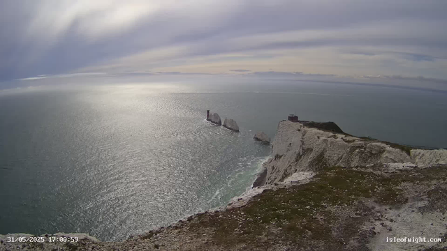 A view of white chalk cliffs overlooking the ocean under a cloudy sky. The cliffs have a grassy top, and there is a pathway along the edge. In the distance, several large rocks are visible in the water near the base of the cliffs. A small building or lighthouse is situated on the cliff's edge. The scene is calm with gentle waves reflecting the overcast sky.