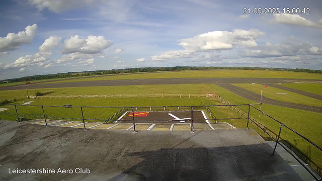 A view from a webcam showing the runway and surrounding grassy airfield under a partly cloudy sky at Leicestershire Aero Club. The date and time are displayed in the upper right corner as 31-05-2025 18:00:42. The foreground shows a tarmac area with a fenced-off helipad marked with a large white "H" and red and white markings. In the bottom left corner, the text "Leicestershire Aero Club" is visible. The horizon reveals open fields and a line of distant trees.
