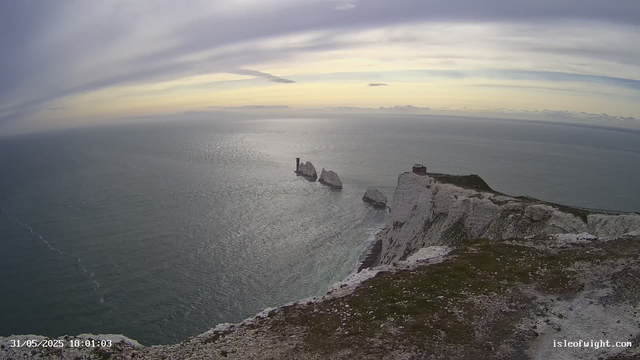 A view of a rugged coastal landscape with white chalk cliffs overlooking the ocean. Several large rock formations or sea stacks rise from the water near the cliffs. A lighthouse or small building is situated on top of the cliffs, and a pathway runs along the edge of the cliff. The sky is partly cloudy with soft, dispersed clouds, and the lighting suggests late evening. The timestamp indicates the date and time as May 31, 2025, at 18:01.