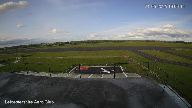 A view from a webcam at Leicestershire Aero Club showing a runway area under a partly cloudy sky. The scene includes a small helipad with a white helicopter silhouette painted on it, surrounded by a grass field and a black safety fence. There is a windsock on the left side, a few taxiway markings, and several runways extending into the distance. The timestamp in the top right corner indicates the date and time as 31st May 2025 at 7:00 PM.