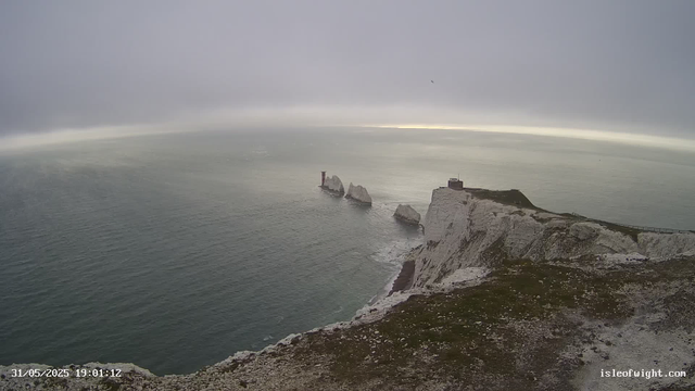 A panoramic view of white chalk cliffs extending into the ocean under a cloudy sky. The cliffs have a rugged edge, with some rocky outcrops near the base. In the distance, there are three prominent rock stacks rising from the water, and a small lighthouse or building is visible on top of the nearest cliff. The ocean appears calm, with gentle waves, and the scene is enveloped in overcast light. The timestamp reads "31/05/2025 19:01:12," and the URL "isleofwight.com" is visible at the bottom right corner.