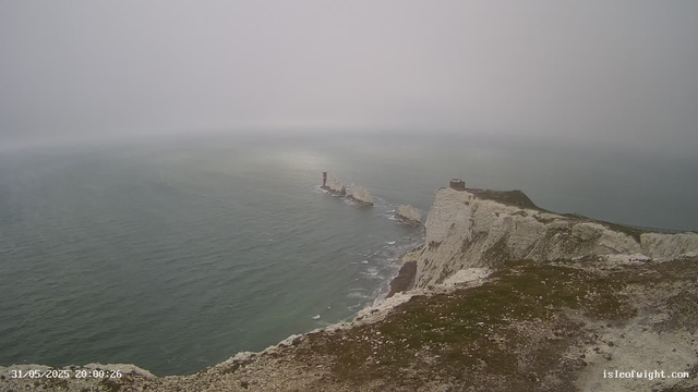 A view of the white chalk cliffs along the coastline, with a lighthouse perched on top of the highest point. The cliffs extend into the sea, which appears calm under a foggy, overcast sky. The image was taken in the evening, as indicated by the timestamp, with a path or railing visible along the edge of the cliff in the foreground.
