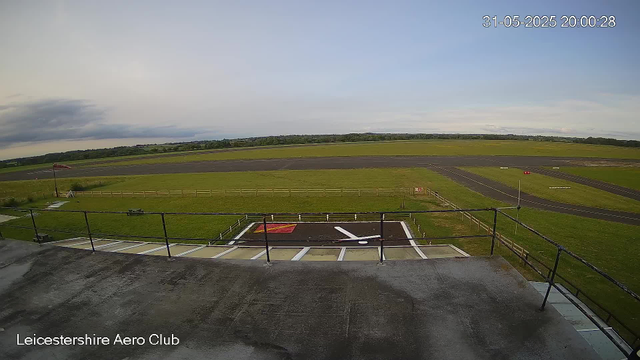 An empty tarmac area with a helicopter landing pad marked with a red square and a white helicopter symbol, surrounded by a black railing. Beyond the tarmac, there is a grassy airfield with a runway and taxiways, with a few distant trees and cloudy sky. In the top right corner, there is a timestamp showing "31-05-2025 20:00:28," and in the bottom left corner, the text reads "Leicestershire Aero Club."