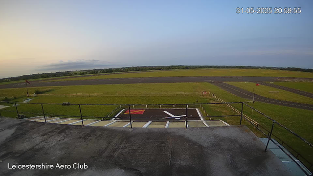 An empty aircraft parking area with a marked helipad, surrounded by a grassy field and bordered by a small wooden fence. In the background, there is a runway with some markings, and beyond that, open fields and distant trees under a partly cloudy sky at sunset. The timestamp in the top right corner shows the date as May 31, 2025, and the time as 20:59:55.