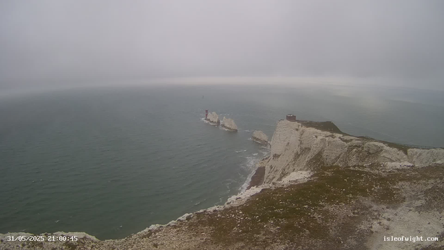 A wide view of white chalk cliffs extending into the ocean on a cloudy day. There are three prominent rock formations near the base of the cliffs, with a lighthouse on one of the distant rocks. The sky is overcast, and the sea appears calm with gentle waves. The coastline curves gently in the distance, and there's a small building at the top of the cliff to the right. The timestamp on the image indicates it was taken on May 31, 2025, at 21:00:45.