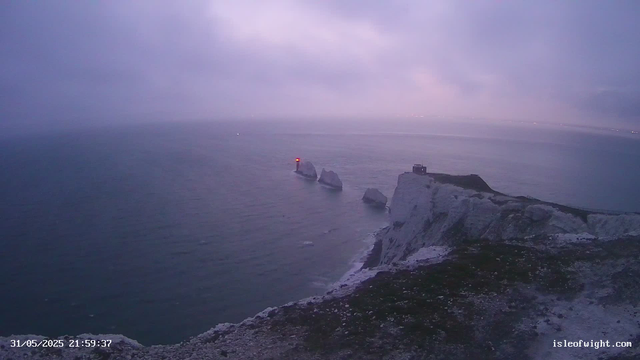 A view of white chalk cliffs along a coast at dusk or dawn, with a few large rocks in the water near the shoreline. There is a small building or lighthouse on the edge of the cliff, and a narrow path or road leading to it. The sky is cloudy with a soft purple hue, and the sea appears calm. In the distance, faint lights are visible along the horizon.
