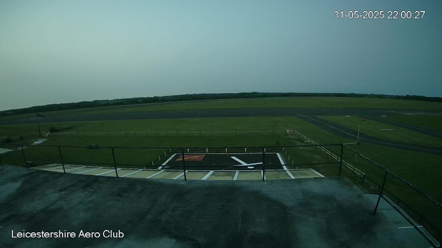 An empty helicopter landing pad with a large black "H" symbol, surrounded by a fence, is visible in the foreground. Beyond the pad, there is a green grassy area with a few small structures, and a winding road extends into the distance. The sky is clear and appears to be around dusk or early evening. The timestamp on the image reads "31-05-2025 22:00:27," and the location is labeled as "Leicestershire Aero Club."
