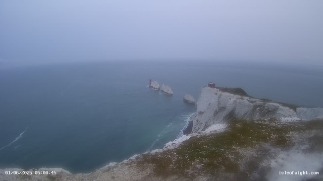 A foggy seascape featuring white chalk cliffs with patches of greenery at the edge. In the distance, there are multiple white chalk stacks rising from the water, with a red and white lighthouse on one of the stacks and a small building on the main cliff. The ocean beneath is calm with gentle waves. The image has a timestamp in the lower left corner and the text "isleofwight.com" in the lower right corner.