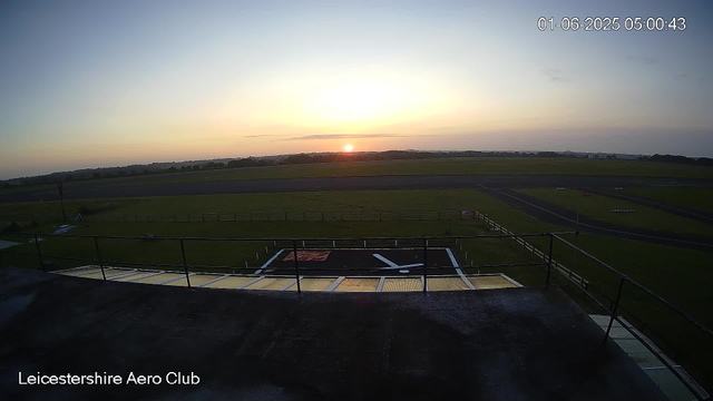A sunrise over a runway and green fields at Leicestershire Aero Club, with a peaceful sky and few clouds. The timestamp shows 05:00 AM on January 6, 2025.