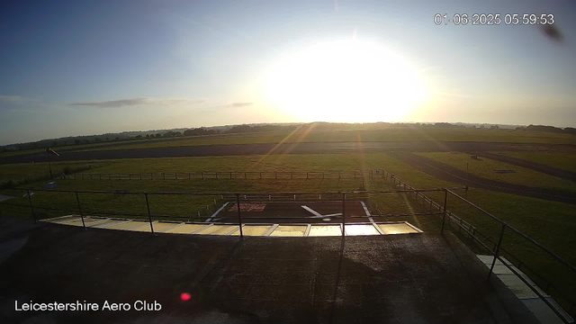 A wide view of a grassy airfield during sunrise, with the sun low on the horizon casting a bright glow. The scene shows part of the Leicesterhire Aero Club; there are safety fences, a helipad, and a windsock on the left. The sky is mostly clear with a few faint clouds. The timestamp in the top right corner indicates the date and time as January 6, 2025, at 5:59 am.