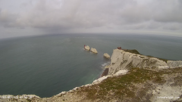 A view of white chalk cliffs with grassy tops extending into the sea, with three rock stacks (the famous chalk stacks known as the Needles) in the water. There is a lighthouse on one of the rock stacks and a small building atop the cliffs. The sky is cloudy and overcast.