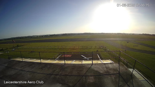 An outdoor view from a webcam at Leicestershire Aero Club showing a grassy field and paved runways in the background, with the sky bright and the sun low on the horizon. The image is timestamped "01-06-2025 07:00:35." A structure with a railing is visible in the foreground.