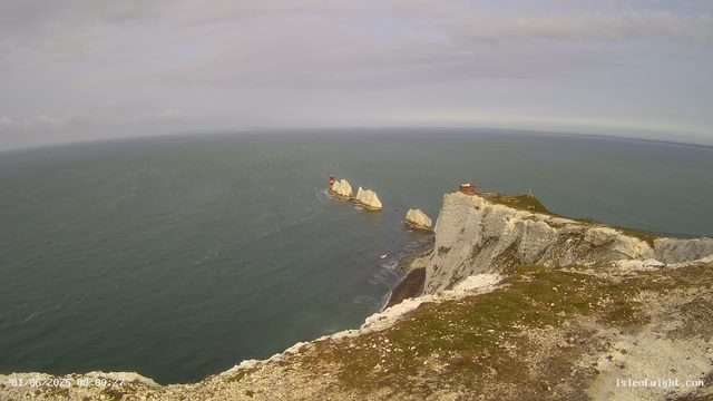 An aerial view of white chalk cliffs with a grassy top, overlooking a vast body of water. A red-and-white striped lighthouse stands on a rock formation in the sea, and a small building is situated on top of the cliffs. The sky is partly cloudy with soft light illuminating the scene.